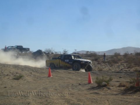 1990 Ford Bronco - yellow and black numbered off-road race truck slides through a dusty desert turn marked by orange cones.