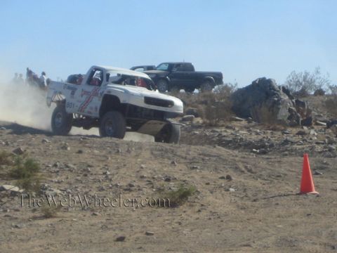 1990 Ford Bronco - white sponsor-decaled off-road race truck powers up a rocky desert grade past an orange cone.