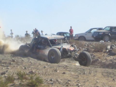 1990 Ford Bronco - open-cockpit off-road race buggy sprays dust through a rocky section as spectators and parked trucks look on.