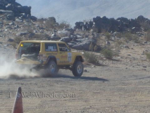 1990 Ford Bronco - yellow Jeep Cherokee-bodied race vehicle turns on a desert course in front of large rock outcrops.