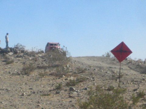 1990 Ford Bronco - red off-road vehicle sits on a rocky desert ridge beside a red diamond course marker as a person stands nearby.