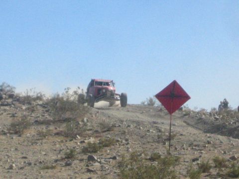 1990 Ford Bronco - off-road vehicle crests a rocky desert hill beside a red diamond course marker under a clear sky.