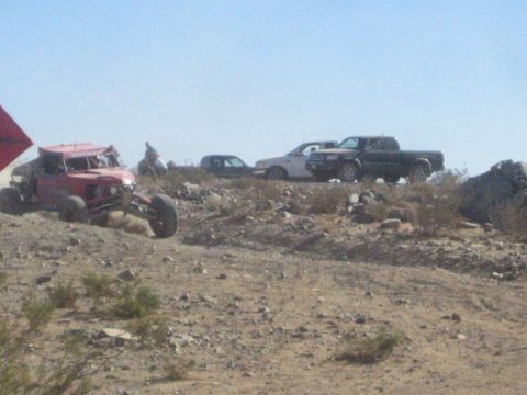 1990 Ford Bronco - red open-wheel off-road buggy sits on a desert ridge with parked spectator trucks lined up behind it.