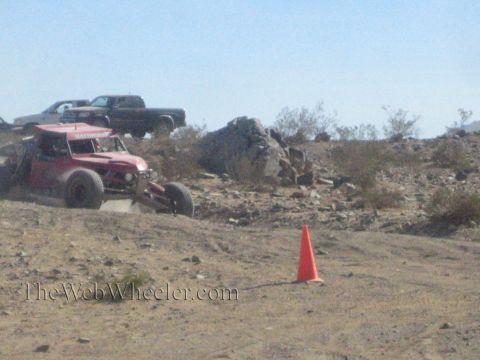 1990 Ford Bronco - red off-road race vehicle sits on a sandy desert course with an orange cone in the foreground and trucks behind.