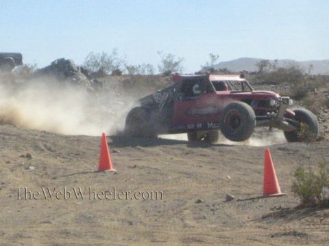 1990 Ford Bronco - pink and purple off-road race buggy drifts through a dusty desert turn between two orange cones.