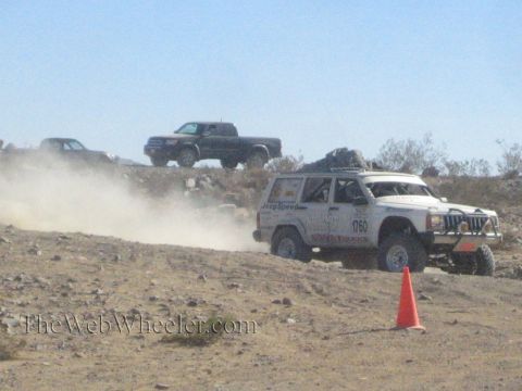 1990 Ford Bronco - white Jeep Cherokee-bodied race vehicle speeds across a rocky desert course trailing dust past an orange cone.