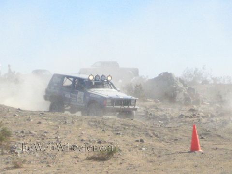 1990 Ford Bronco - blue Jeep Cherokee-bodied race vehicle with a roof light bar charges through heavy desert dust.