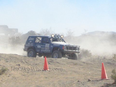 1990 Ford Bronco - blue Jeep Cherokee-bodied race vehicle with a roof light bar slides through a dusty desert turn near orange cones.