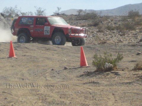 1990 Ford Bronco - red Jeep Cherokee-bodied race vehicle accelerates across a sandy desert course between orange cones.