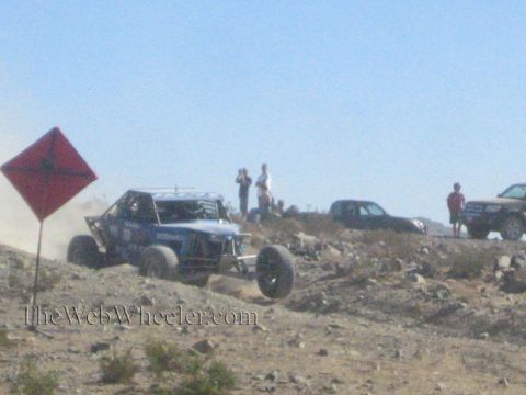 1990 Ford Bronco - blue off-road race buggy climbs a rocky rise beside a red diamond marker as spectators watch from above.