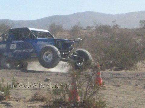 1990 Ford Bronco - blue numbered off-road race truck catches air over a desert jump, trailing dust against a mountain backdrop.