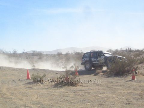 1990 Ford Bronco - black Jeep Cherokee-bodied race vehicle rounds a dusty desert turn marked by orange cones against distant mountains.