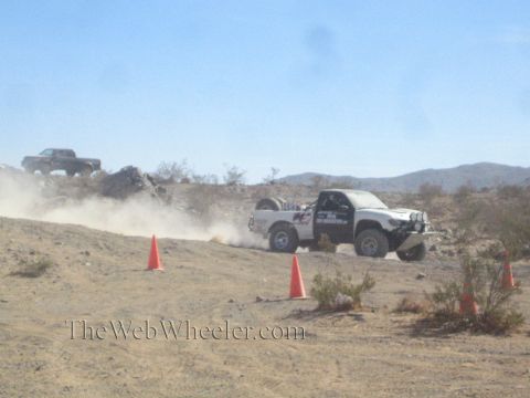 1990 Ford Bronco - white off-road race truck speeds across a desert course between orange cones, raising dust against mountains.