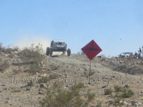 1990 Ford Bronco - off-road race buggy crosses a rocky desert rise beside a red diamond course marker under a clear sky.