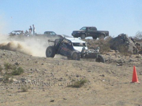 1990 Ford Bronco - open-cockpit off-road race buggy slides sideways on a desert berm as spectators and trucks watch from above.