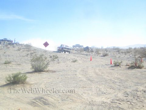 1990 Ford Bronco - white off-road race truck crests a dusty desert rise past a course marker sign and traffic cones under a clear sky.