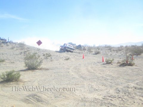 1990 Ford Bronco - off-road truck throws up a large dust cloud as it crosses a desert ridge beside a diamond-shaped course sign.