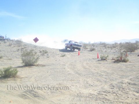 1990 Ford Bronco - white pickup-style race truck speeds across rocky desert ground, trailing dust between orange marker cones.