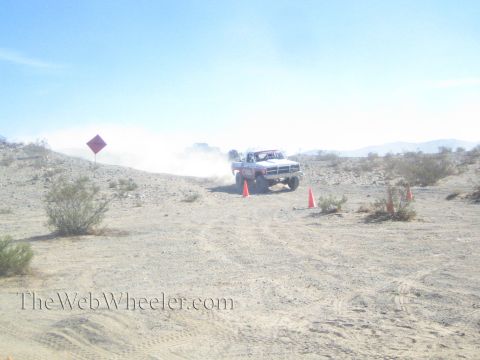 1990 Ford Bronco - lifted white off-road truck with a roll cage climbs a sandy desert hill, raising dust beside a course sign and cones.