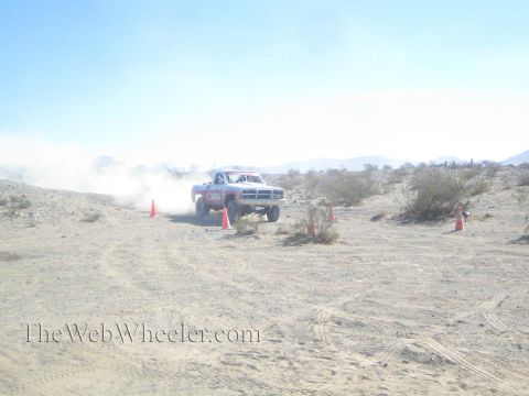 1990 Ford Bronco - off-road race truck catches air over uneven desert ground, kicking up a dense dust cloud near orange cones.