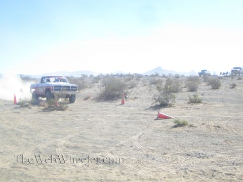 1990 Ford Bronco - white off-road truck lifts its front wheels off the ground in open desert, trailing dust with mountains behind.