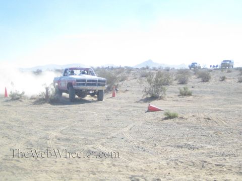 1990 Ford Bronco - white Dodge Ram off-road truck charges head-on across the desert, raising dust beside orange marker cones.