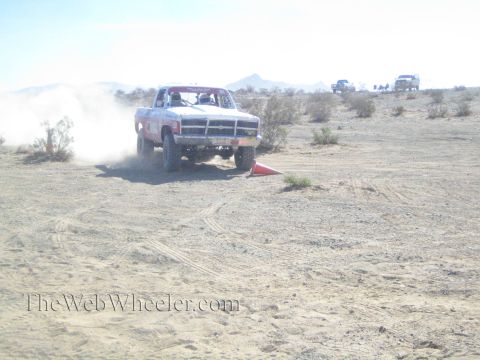 1990 Ford Bronco - white Dodge off-road race truck drives front-on over desert terrain across a red mat, trailing a dust cloud.