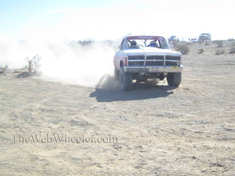 1990 Ford Bronco - white off-road race truck drives toward the camera shrouded in thick dust on a rough desert track.