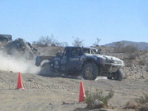 1990 Ford Bronco - black off-road race truck numbered 53 powers across rocky desert ground, raising dust between orange cones.