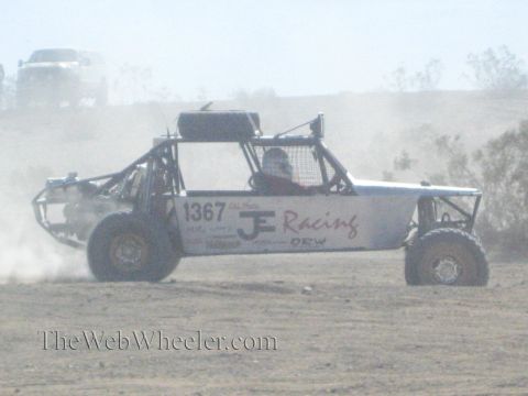 1990 Ford Bronco - tube-frame off-road buggy marked 1367 JE Racing speeds in profile across a dusty desert flat.