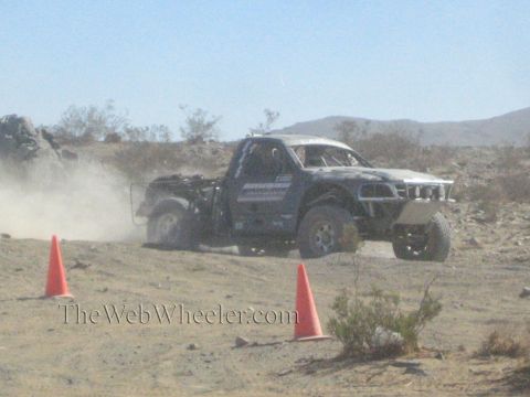 1990 Ford Bronco - silver-and-black off-road race truck drives along a desert track past orange cones, kicking up dust.