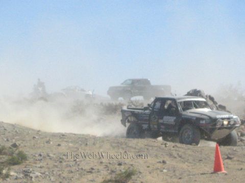 1990 Ford Bronco - off-road race truck pushes through thick dust on a desert course while a dark truck follows in the haze.