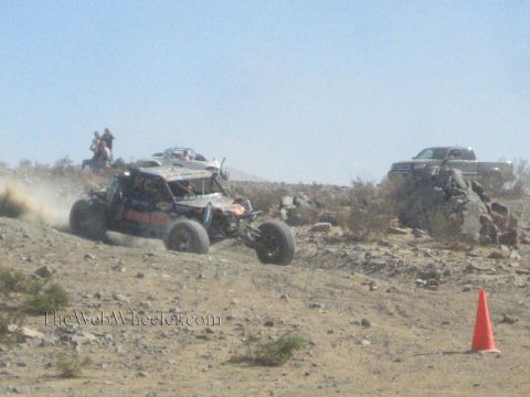 1990 Ford Bronco - white tube-frame off-road buggy crosses a rocky desert slope as spectators and a chase truck watch nearby.