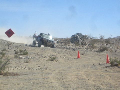 1990 Ford Bronco - lifted off-road truck with spotters aboard climbs a sandy desert hill beside a course warning sign and cones.