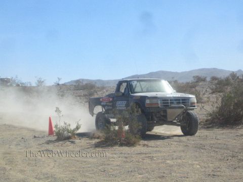 1990 Ford Bronco - dark Ford off-road race truck drives toward the camera across desert ground, throwing up dust beside a cone.