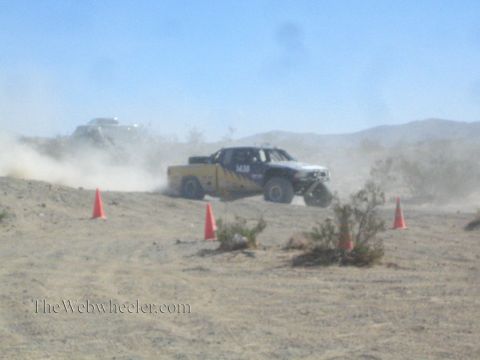 1990 Ford Bronco - yellow-and-white off-road race truck slides across a desert rise in a cloud of dust between orange cones.