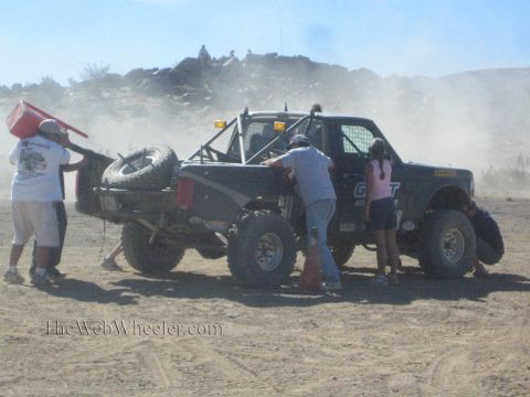 1990 Ford Bronco - pit crew tends to a black off-road race truck, refueling and checking the wheel in a dusty desert staging area.