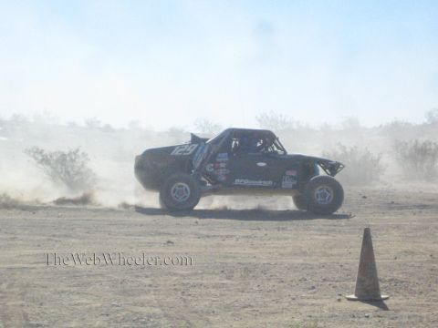1990 Ford Bronco - black off-road race truck numbered 129 slides sideways across the desert in a thick dust cloud near a cone.