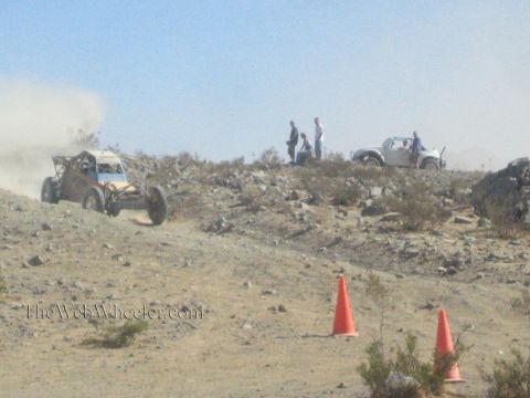 1990 Ford Bronco - tube-frame off-road buggy climbs a rocky desert slope while spectators stand near a parked vehicle above.