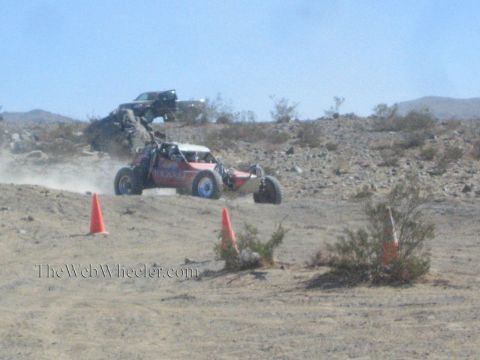 1990 Ford Bronco - red-and-white tube-frame off-road buggy drives across desert ground past orange cones, raising dust.