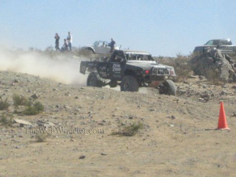 1990 Ford Bronco - black off-road race truck powers up a rocky desert hill, trailing dust as spectators watch from above.