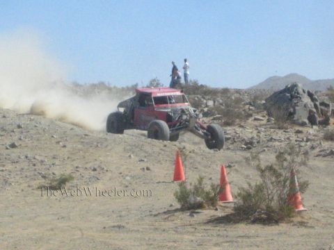 1990 Ford Bronco - red tube-frame off-road buggy catches air over a desert rise near orange cones as spectators look on.