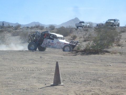 1990 Ford Bronco - white tube-frame off-road buggy with an exposed rear engine slides across a desert flat past a tall cone.