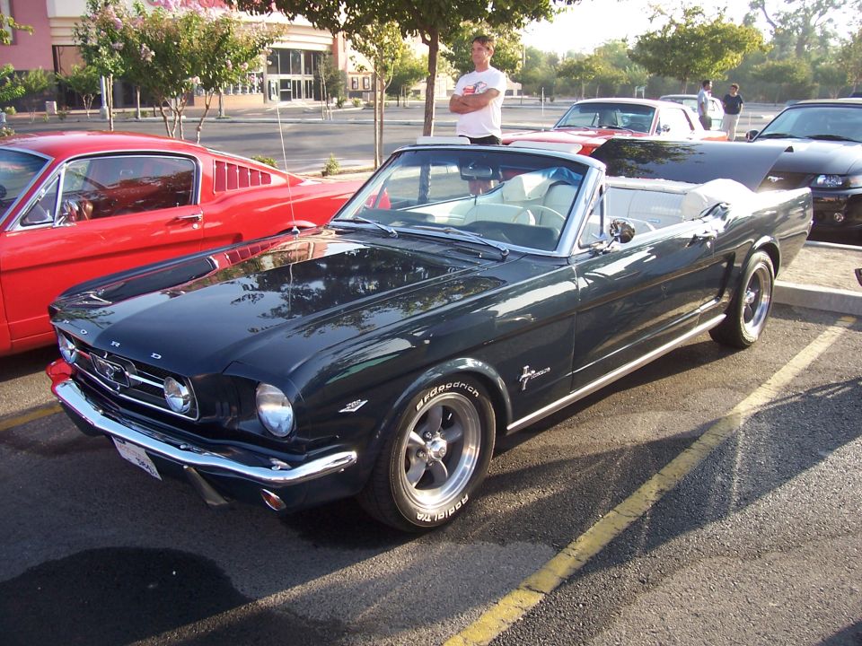 Dark green convertible classic car parked next to a red coupe. Guy in white shirt stands behind it
