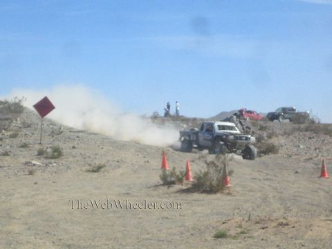 1990 Ford Bronco - off-road truck throws up a dust cloud cresting a desert hill beside a marker sign, cones, and distant spectators.