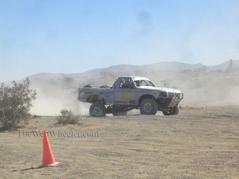 1990 Ford Bronco - white off-road race truck marked 0310 races across a dusty desert track with mountains in the background.