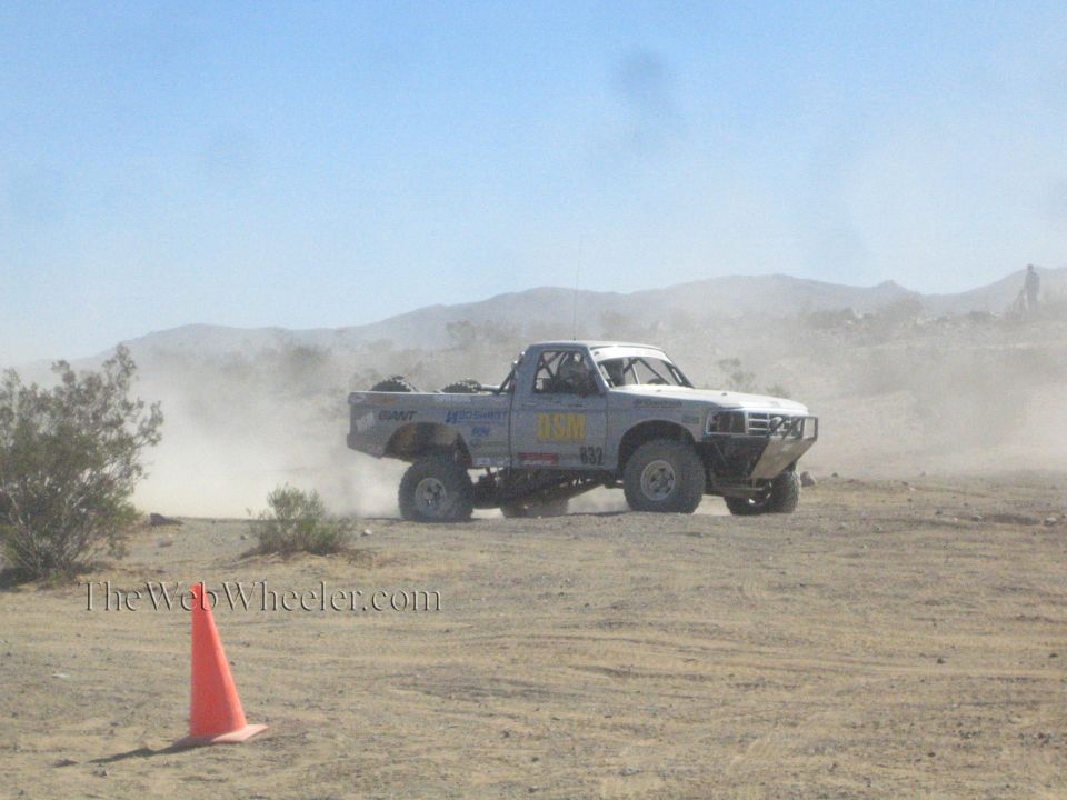 White off-road race truck number 0310 kicking up dust on a desert course past a traffic cone