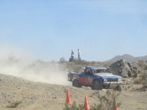1990 Ford Bronco - blue and red prerunner-style race truck speeds along a rocky desert ridge as spectators watch from above.