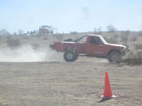 1990 Ford Bronco - orange-red off-road race truck runs a dusty desert course past a traffic cone with a white truck behind.