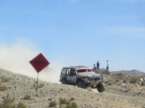1990 Ford Bronco - modified lifted SUV race vehicle climbs a rocky desert grade past a red diamond course marker as spectators watch.